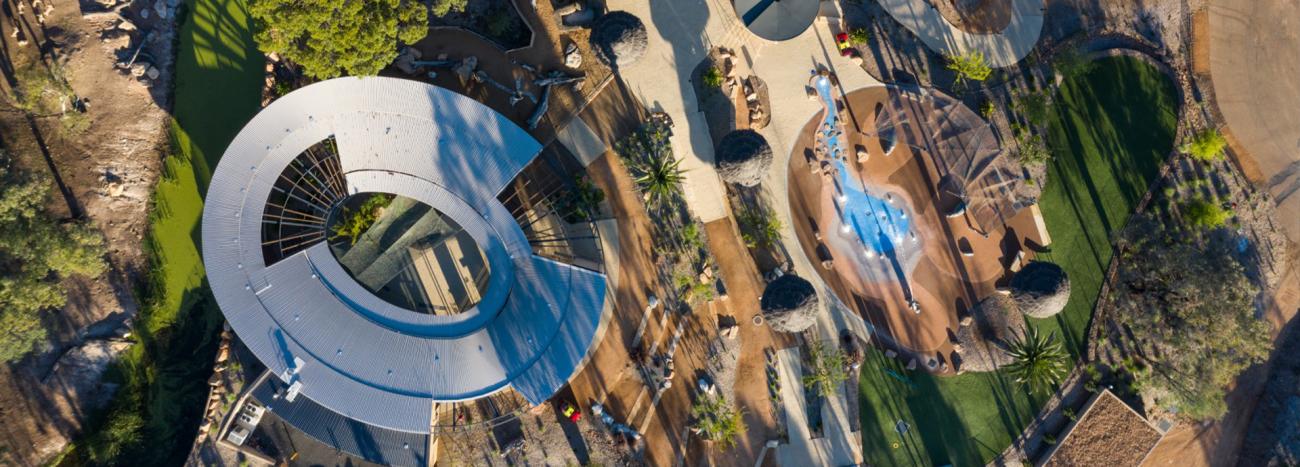 Aerial view of a modern architecture project featuring a curved steel roof, landscaped pathways, greenery, and a nature-inspired playground. 