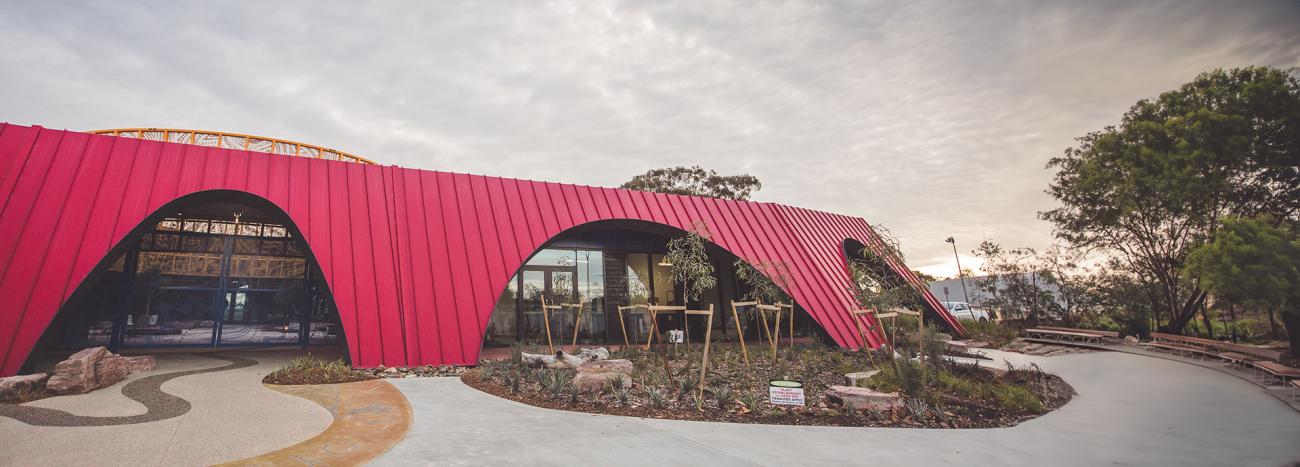 A modern building featuring a bold red facade made of KLIP-LOK 700 HI-STRENGTH® metal sheeting, with distinctive curved archways. Large glass windows and doors reflect the surrounding trees and landscape. 